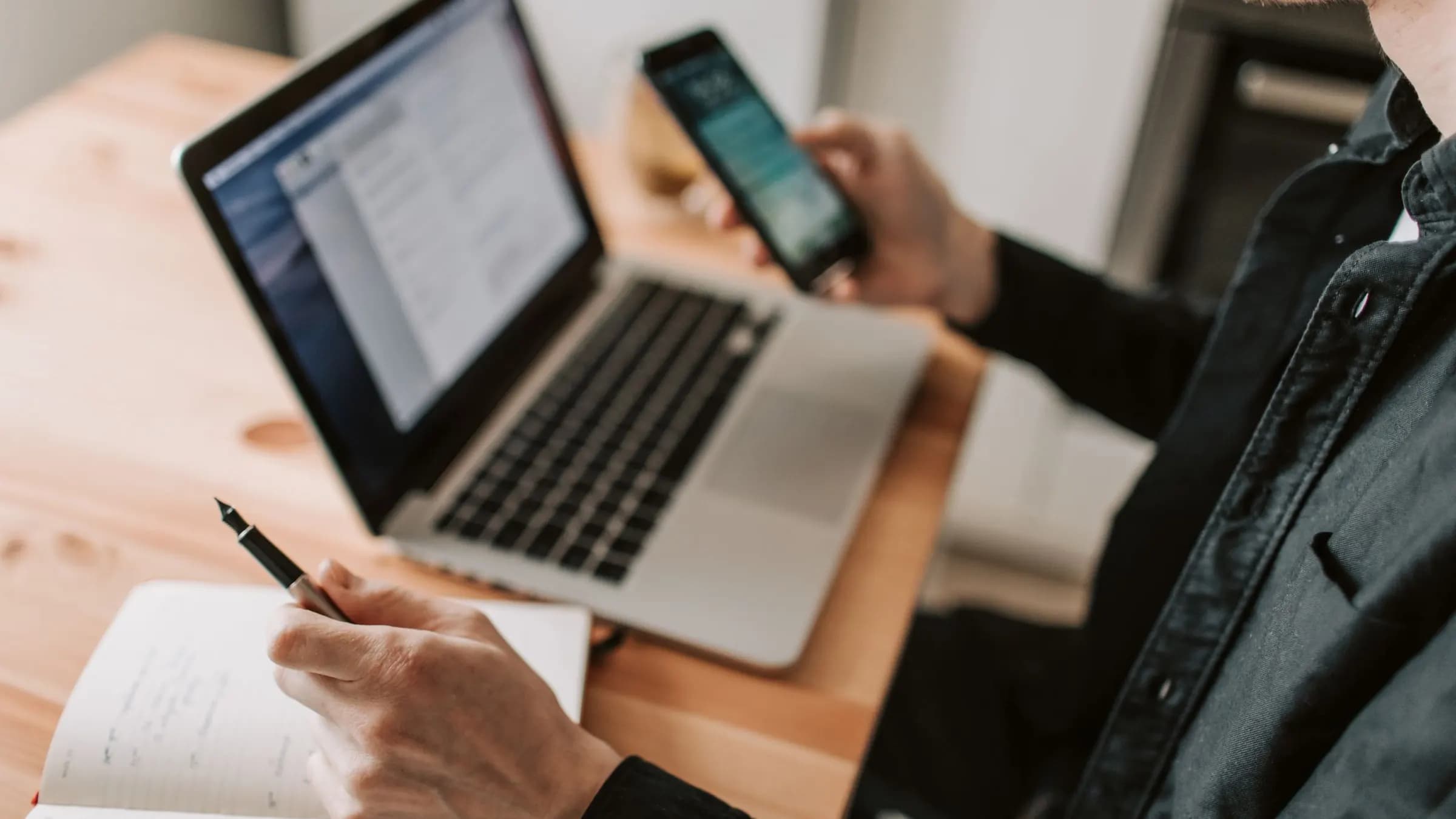Someone using a phone beside an open laptop and a notebook on a desk.